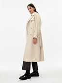 side view of a woman wearing a beige double breasted cashmere trench coat with belt  over a sweater and dark pants on a gray background #color_beige