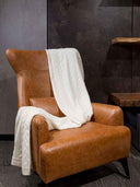 A cable knit blanket in beige color draped over a brown leather armchair. #color_white