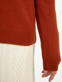 close up of a red-orange cardigan sleeve and white knitted pants on a light gray background #color_sugar-almond